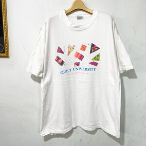 以下が含まれることがあります： 白い半袖Tシャツ。胸の中央に、様々な色や柄の布製三角形のグラフィックと、青い文字で「QUILT UNIVERSITY」というテキスト、その下に小さな文字がプリントされています。