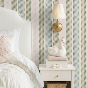 May include: A bedroom scene featuring a white upholstered headboard, bed with white bedding, and a round pink pillow. A white nightstand holds a lamp, a swan figurine, and a woven basket. The wallpaper has vertical stripes in pink, green, and white.