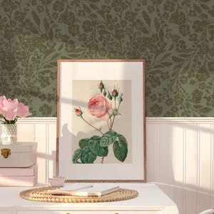 Framed botanical art with a rose on a white desk, next to pink flower vase and stacked boxes. Green floral wallpaper adds a vintage touch. Cozy ambiance.
