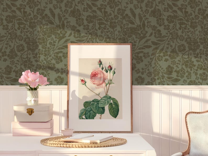 Framed botanical art with a rose on a white desk, next to pink flower vase and stacked boxes. Green floral wallpaper adds a vintage touch. Cozy ambiance.