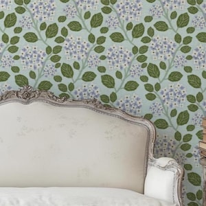 May include: A light blue floral wallpaper with a repeating pattern of green leaves and clusters of purple flowers. A white upholstered headboard and a stack of books on a small table are in the foreground.