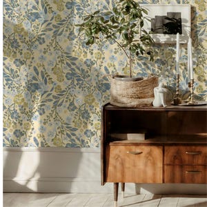 May include: A room interior featuring floral wallpaper in shades of blue, green, and cream. A wooden cabinet with drawers and a woven basket holding a plant are in the foreground. Decorative candles and a framed artwork complete the scene.