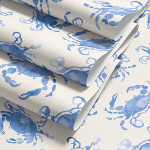 May include: Rolls of wallpaper featuring a repeating pattern of blue watercolor crabs on a cream background. The crab design is detailed and artistic, with a coastal or nautical theme. The rolls are partially unrolled, showing the pattern.