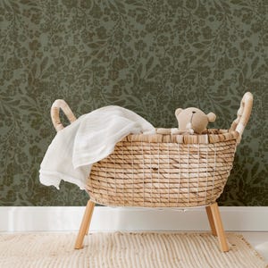 A wicker baby bassinet with a teddy bear and white blanket sits on a textured rug. The backdrop features dark green floral wallpaper, creating a cozy, serene atmosphere