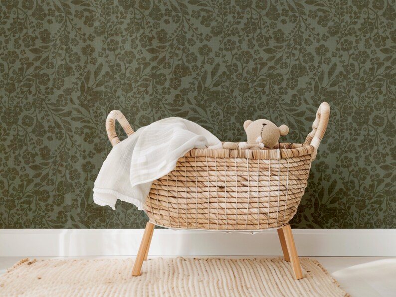 A wicker baby bassinet with a teddy bear and white blanket sits on a textured rug. The backdrop features dark green floral wallpaper, creating a cozy, serene atmosphere