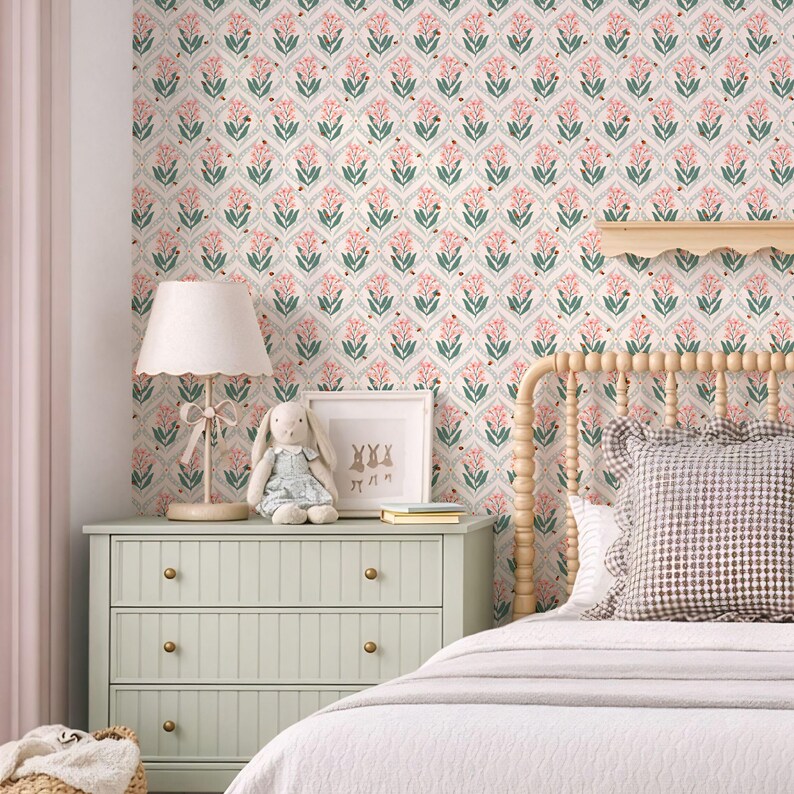 Wallpaper featuring a soft floral pattern in muted pink, sage green, and cream tones with a delicate repeating botanical design. The scene shows a cozy nursery or child's bedroom with a wooden spindle bed, gingham pillows, a light green dresser, plush bunny toys, a scalloped lampshade, and a neutral area rug.