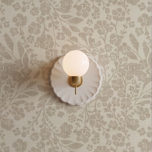 May include: A wall sconce featuring a circular, scalloped white backplate and a central brass-toned fixture holding a spherical, frosted white glass globe light bulb that emits a soft, warm glow against the wall surface.