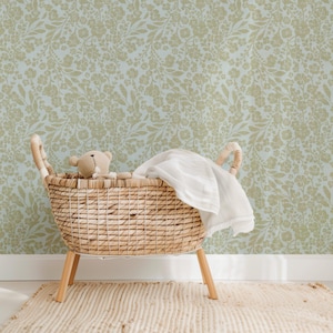 K&ouml;nnte beinhalten: Ein Weidenkorb-Babybett mit einer wei&szlig;en Decke und einem braunen Teddyb&auml;ren im Inneren. Das Babybett steht auf einem hellbraunen Teppich vor einer Wand mit einem hellblauen und braunen Blumenmuster.