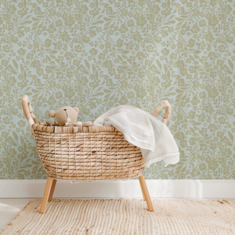 K&ouml;nnte beinhalten: Ein Weidenkorb-Babybett mit einer wei&szlig;en Decke und einem braunen Teddyb&auml;ren im Inneren. Das Babybett steht auf einem hellbraunen Teppich vor einer Wand mit einem hellblauen und braunen Blumenmuster.