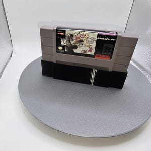 May include: A gray Super Nintendo video game cartridge, labeled "Chrono Trigger" by SquareSoft, is displayed on a black stand. The cartridge features colorful artwork and the rating "KA". The game is set on a gray circular surface.