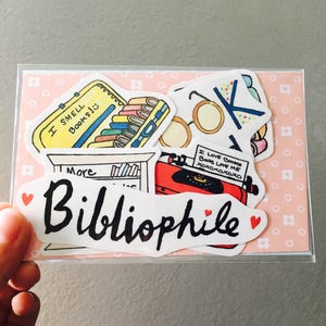 May include: A collection of colorful stickers with a book theme. The stickers include a yellow suitcase with books, a typewriter, and the word "Bibliophile". The text "I SMELL Books!" and "I LOVE Books Books LOVE ME xoxoxoxoxo" are also visible.