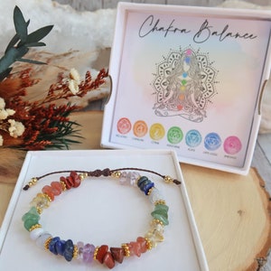 May include: A chakra bracelet featuring seven different coloured stones, including red, orange, yellow, green, blue, indigo, and violet. The bracelet is made with a brown cord and gold beads. The bracelet is displayed in a white box with the text "Chakra Balance" on the lid.