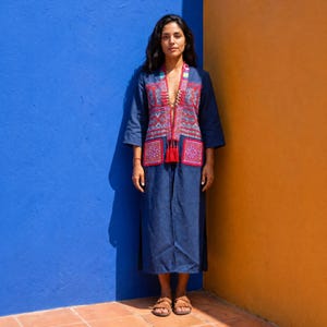 Può includere: Un cappotto lungo a maniche lunghe blu scuro caratterizzato da un profondo scollo a V con intricati ricami geometrici rossi, bianchi e blu sui pannelli frontali e due grandi tasche quadrate, rifinito con un dettaglio a nappa rossa sul petto.