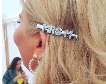 Könnte beinhalten: Eine silberfarbene Haarspange aus Metall mit dem Schriftzug MRS M, der aus klaren, funkelnden Strasssteinen besteht. Das Accessoire ist horizontal im blonden Haar befestigt und setzt einen dekorativen Akzent, der durch die Oberfläche das Licht reflektiert.