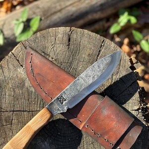 以下が含まれることがあります： 木製の柄と革製の鞘が付いた手作りのナイフ。 刃はテクスチャード加工が施され、装飾的なエンブレムが付いています。 鞘は茶色でステッチされています。 ナイフは木製の表面に置かれています。