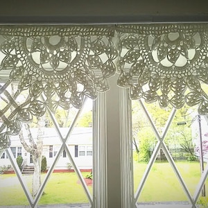 May include: White crocheted lace valance with a scalloped edge hangs in front of a window with diamond-shaped panes. The window looks out onto a green lawn and trees.