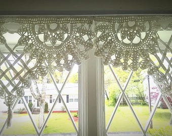 May include: White crocheted lace valance with a scalloped edge hangs in front of a window with diamond-shaped panes. The window looks out onto a green lawn and trees.