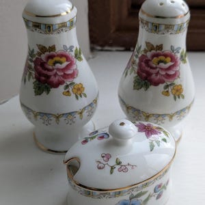 May include: A white porcelain salt and pepper shaker set and a small lidded dish. The set features a floral design with red, pink, yellow, and blue flowers, accented with gold trim. The salt and pepper shakers are approximately 4 inches tall.