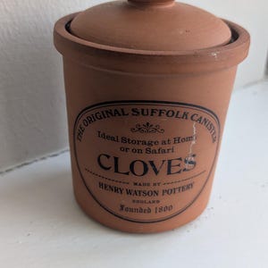 May include: A terracotta canister with a lid and a round knob. The canister has a label that reads "CLOVES" and "HENRY WATSON POTTERY ENGLAND Founded 1800". The canister is designed for storage.