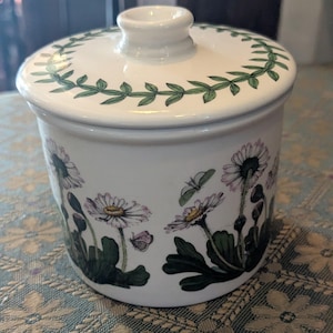 May include: A white ceramic jar with a lid, decorated with a floral pattern of daisies and butterflies. The lid has a green leaf border. The jar is cylindrical and has a small handle on top of the lid.