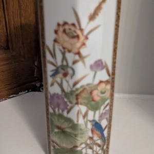 Puede incluir: Un jarrón rectangular blanco con un diseño floral y de pájaros. El jarrón presenta un borde dorado y está decorado con flores coloridas, hojas verdes y pájaros azules. El jarrón es un objeto decorativo.