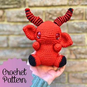 May include: Crochet pattern for a red amigurumi devil with black horns and eyes. The devil is holding a pink sign that says "Crochet Pattern".