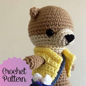 May include: Crocheted otter amigurumi toy wearing a yellow vest and a blue satchel. The otter has brown fur, black eyes, and a black nose. The text "Crochet Pattern" is visible in the image.