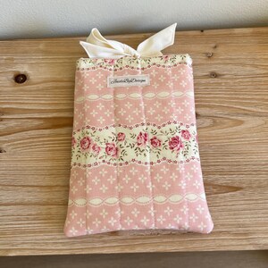 May include: A pink floral fabric book cover with a cream-colored bow. The cover features a repeating pattern of pink roses and white lace-like details. The text "InsideByDesign" is printed on a small label.