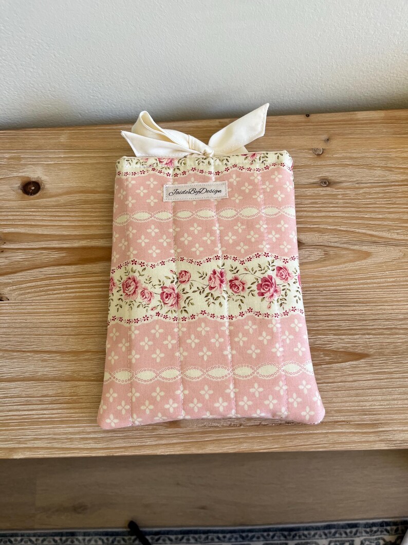 May include: A pink floral fabric book cover with a cream-colored bow. The cover features a repeating pattern of pink roses and white lace-like details. The text "InsideByDesign" is printed on a small label.