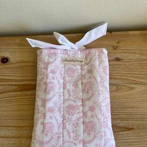 May include: A rectangular, quilted fabric item with a white background and a pink floral pattern. The item is tied with a white ribbon. The text "Inside By Design" is printed on a small label.