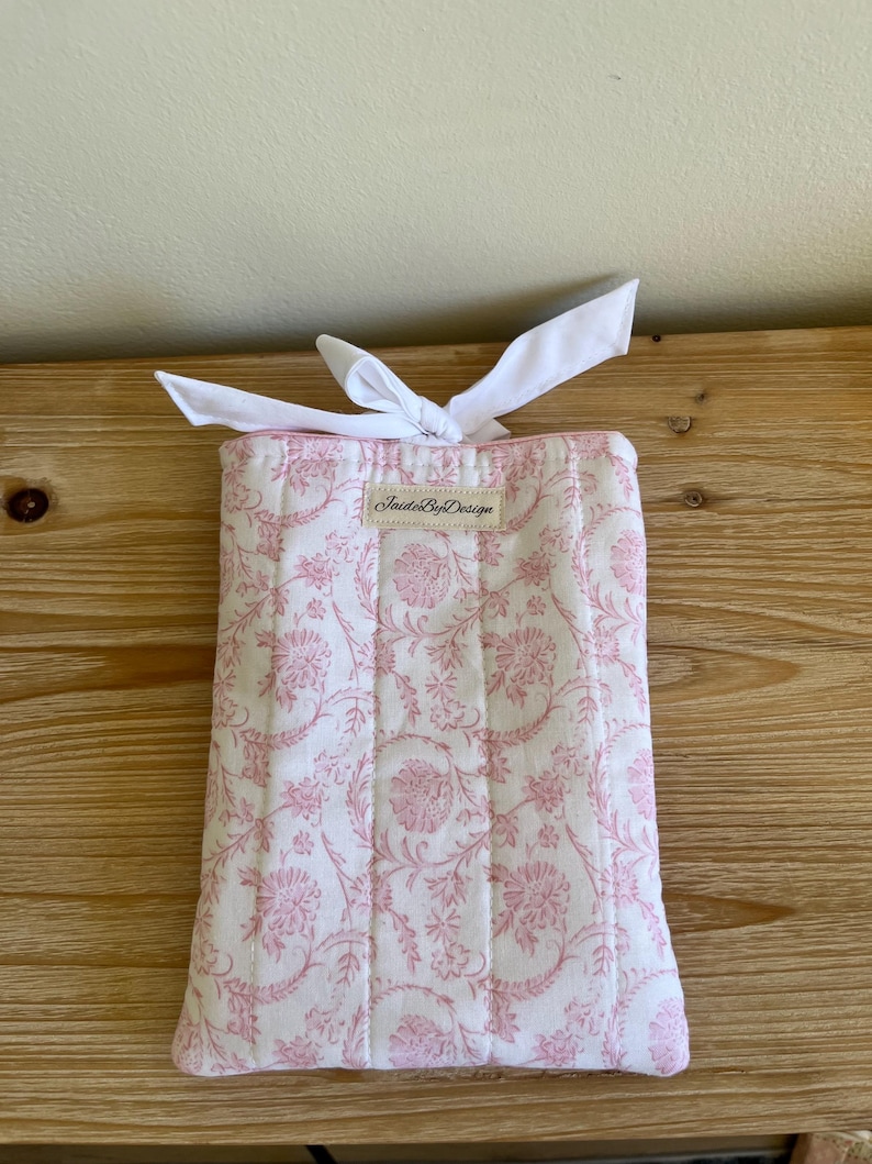 May include: A rectangular, quilted fabric item with a white background and a pink floral pattern. The item is tied with a white ribbon. The text "Inside By Design" is printed on a small label.