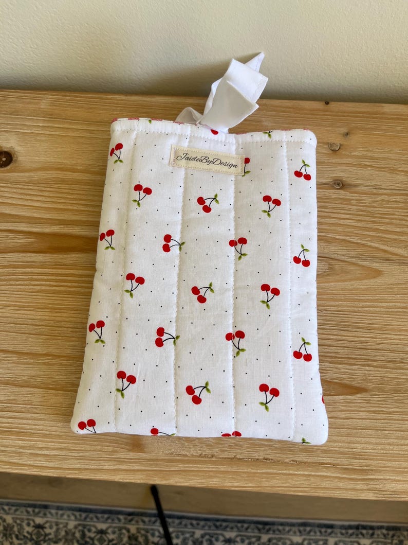 May include: White quilted fabric case with a cherry pattern. The case features red cherries with green stems and leaves, scattered across a white background with small black dots. The case has a white ribbon and the text "JaideByDesign".