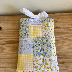 May include: A quilted fabric item with a patchwork design. The design includes yellow gingham, floral patterns in blue and yellow, and a blue floral print. A white ribbon is tied at the top. The text "JaideByDesign" is visible.