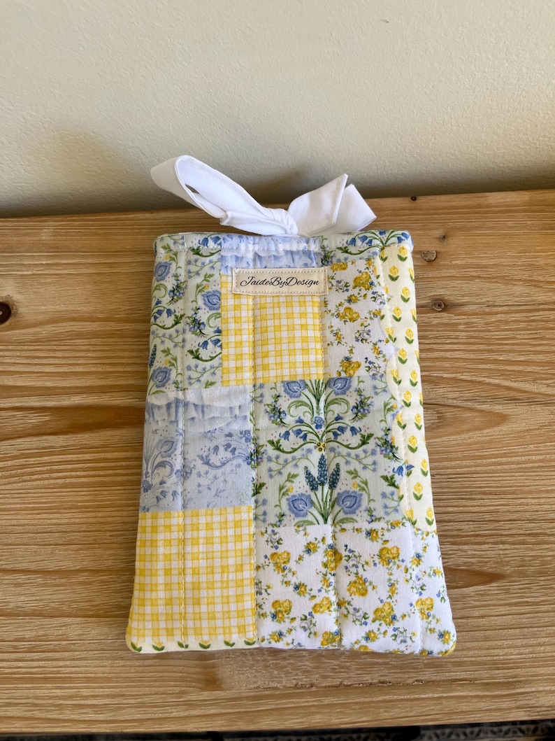 May include: A quilted fabric item with a patchwork design. The design includes yellow gingham, floral patterns in blue and yellow, and a blue floral print. A white ribbon is tied at the top. The text "JaideByDesign" is visible.