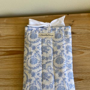 May include: A quilted fabric case with a blue floral pattern on a white background. The case is topped with a white bow. The label "JaideByDesign" is visible on the front of the case. The case is rectangular and appears to be a protective sleeve.