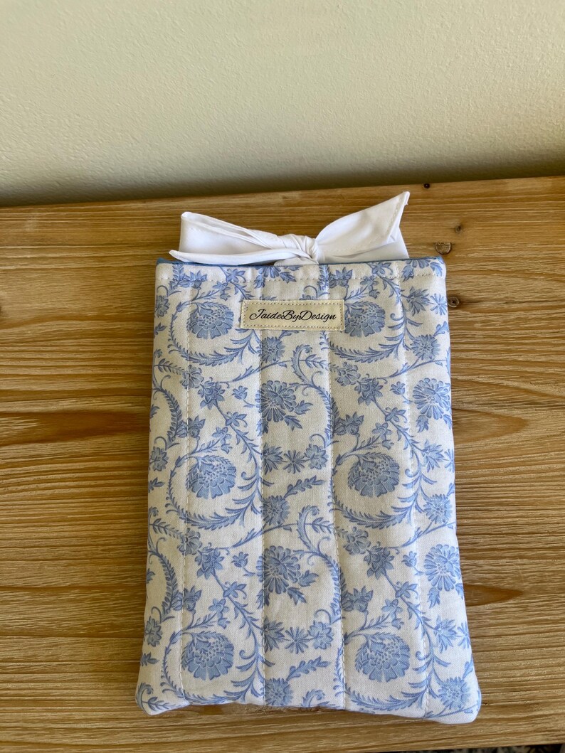May include: A quilted fabric case with a blue floral pattern on a white background. The case is topped with a white bow. The label "JaideByDesign" is visible on the front of the case. The case is rectangular and appears to be a protective sleeve.