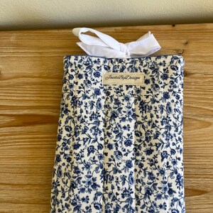 May include: A rectangular, quilted fabric item with a white and blue floral pattern. A white bow is tied at the top. The label "JaideByDesign" is visible on the front.