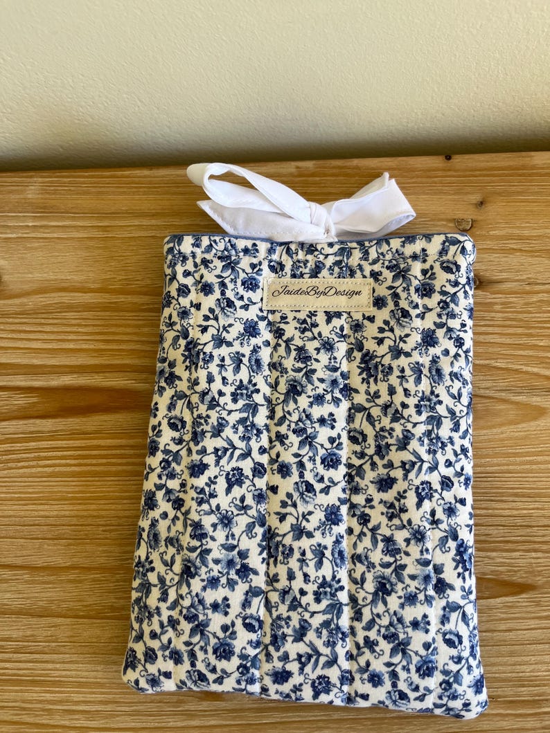 May include: A rectangular, quilted fabric item with a white and blue floral pattern. A white bow is tied at the top. The label "JaideByDesign" is visible on the front.