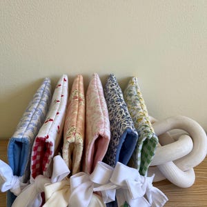 May include: A collection of fabric pot holders in various floral and patterned designs, including red gingham, blue toile, and floral prints. Each pot holder is tied with a white ribbon bow. A white wooden decorative chain is on the right.