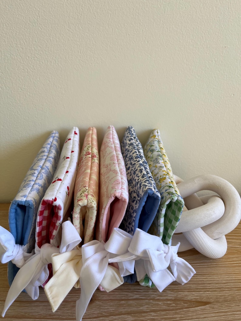 May include: A collection of fabric pot holders in various floral and patterned designs, including red gingham, blue toile, and floral prints. Each pot holder is tied with a white ribbon bow. A white wooden decorative chain is on the right.
