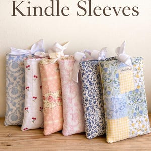 May include: A collection of quilted Kindle sleeves in various floral and patterned fabrics, each tied with a white ribbon. The sleeves are displayed on a wooden surface, with text that reads "Quilted Kindle Sleeves" and "Handmade • Fits Paperwhite & More".