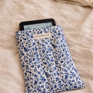 May include: A blue and white floral print e-reader sleeve with an e-reader inside. The sleeve has a label that says "JaideByDesign". The text "Handmade & designed to actually fit" is at the bottom.