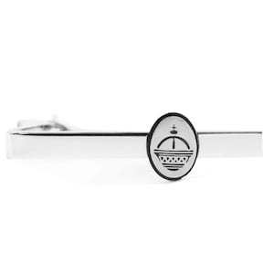 May include: A silver-toned tie clip with an oval emblem featuring a crown design. The crown is black and white, and the clip has a polished, reflective surface. The tie clip is a formal accessory.