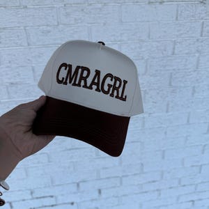 May include: A white and brown baseball cap with the text "CMRAGRL" embroidered on the front. The cap has a brown brim and a white crown. The hat is held up in front of a white brick wall.