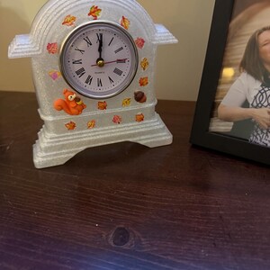 May include: A decorative quartz clock with a white, glittery finish. The clock face has Roman numerals and gold hands. The clock is adorned with colorful autumn leaves and a small orange squirrel figurine. A black framed photo is in the background.