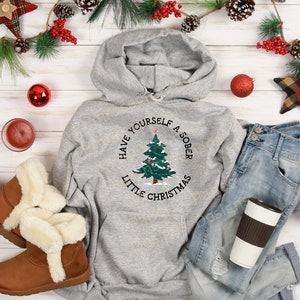 May include: A gray hooded sweatshirt with a Christmas tree design and the text "Have yourself a sober little Christmas".