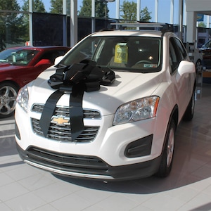May include: A white Chevrolet Trax SUV with a large black bow on the hood. The car is parked in a showroom with a red Chevrolet Camaro in the background.