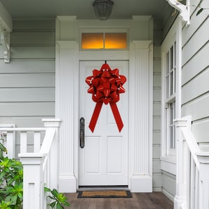 May include: A large red bow with a satin ribbon is attached to a white front door. The bow is a festive decoration for a home.