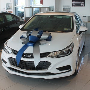May include: A white Chevrolet sedan with a large blue and silver bow on the hood. The car is in a showroom setting.