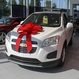 May include: A white SUV with a red bow on the hood. The car has a black grill and headlights. The car is parked in a showroom.